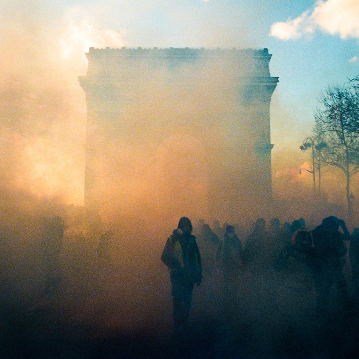 A crowd of people is shrouded in yellow smoke in front of the Arc de Triomphe.