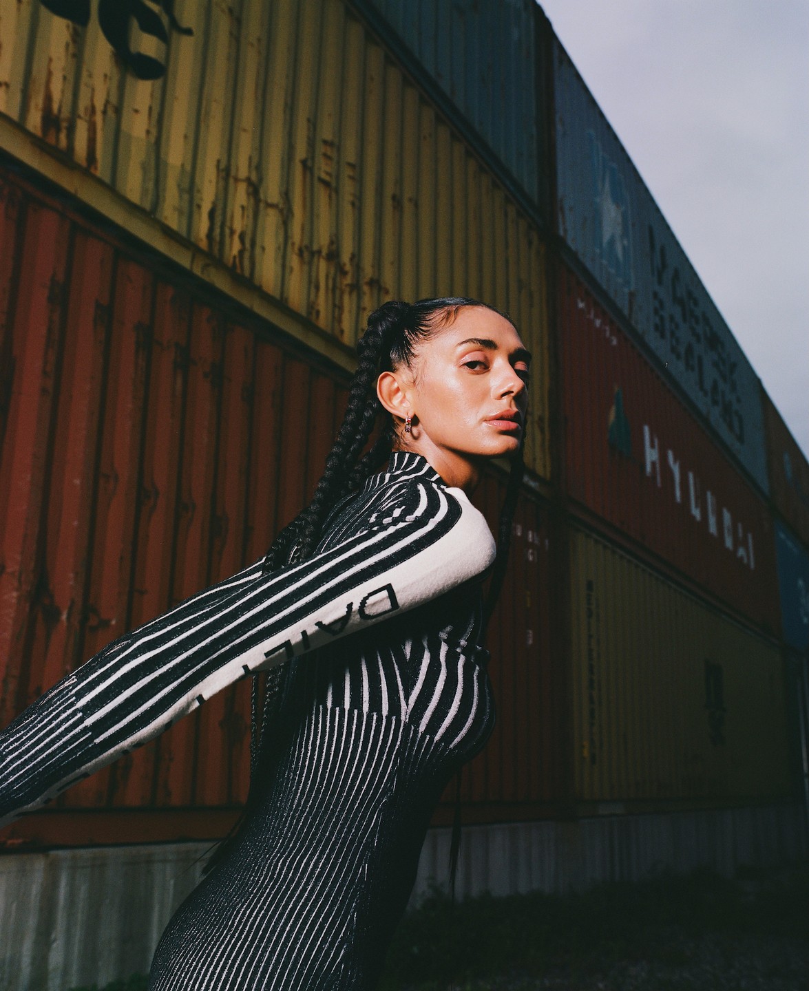 A woman with braided hair and a striped outfit poses in front of shipping containers.
