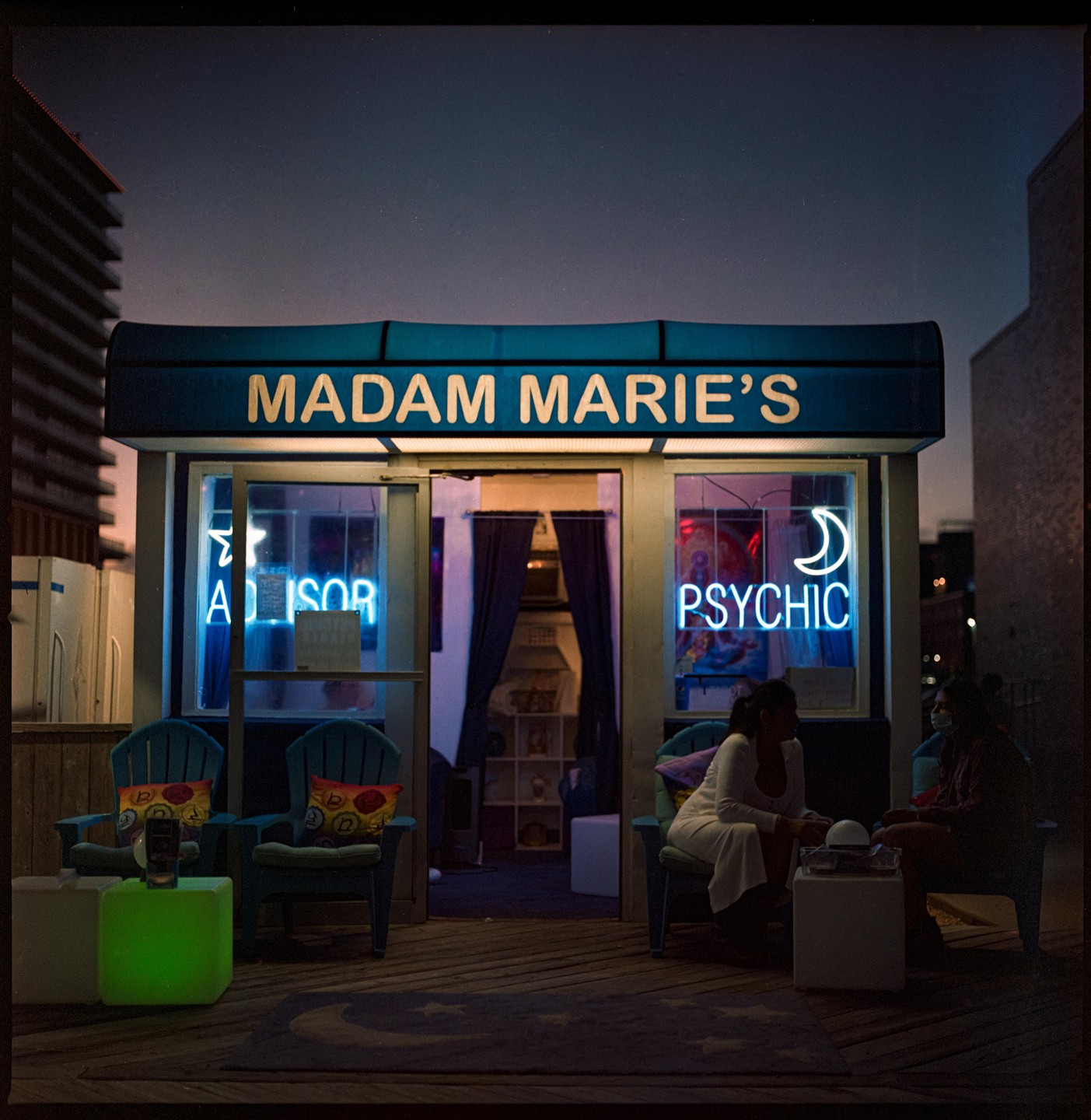 Two women sit outside Madam Marie's psychic shop at night.