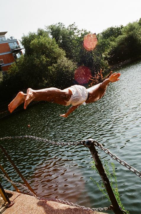 A person dives into a body of water, with trees and a building visible in the background.