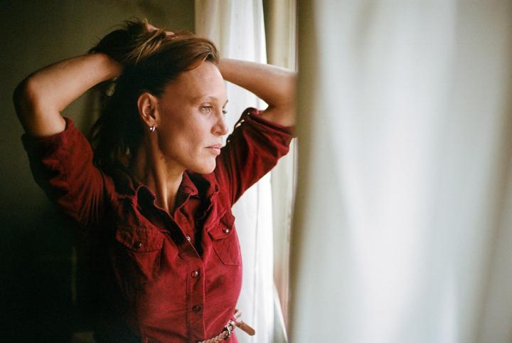 A woman in a red shirt looks out a window, with her hands in her hair.
