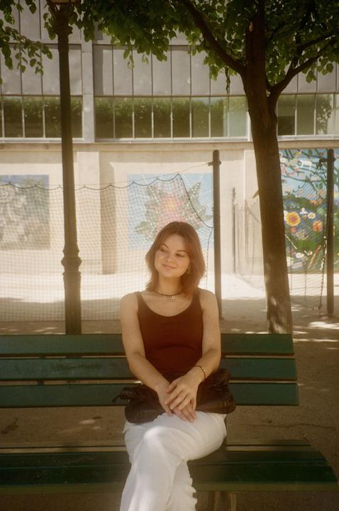A woman sits on a green park bench under a tree, with a building and a colorful mural in the background.