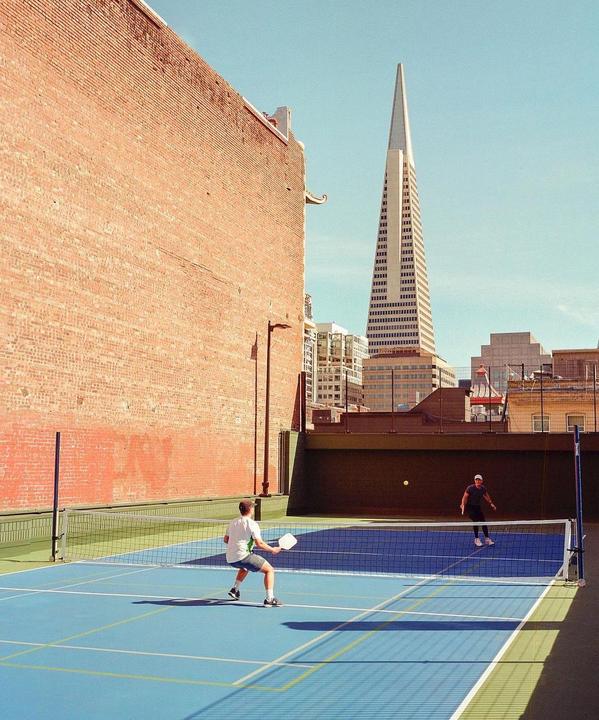 Two men play pickleball on a blue court next to a tall brick building with a skyscraper in the background.