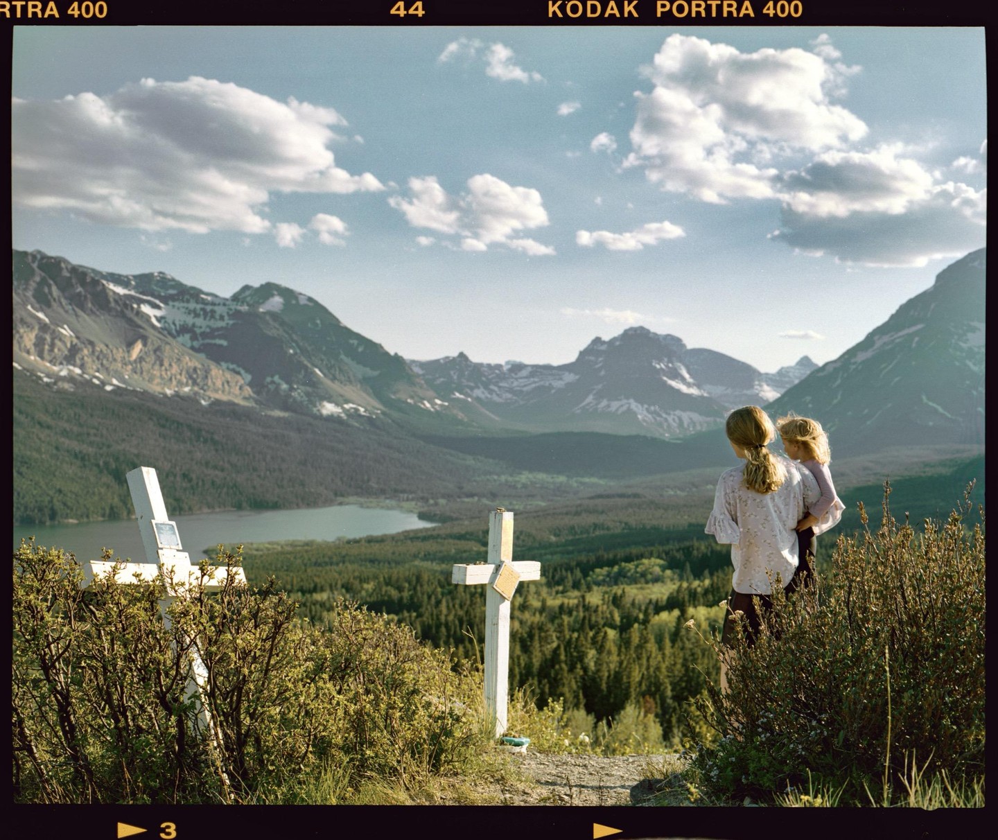 Two girls stand on a hill overlooking a lake and mountains, with two white crosses in the foreground.