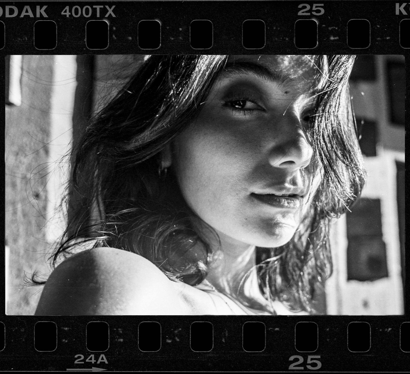 A woman with dark hair looks towards the viewer, with sunlight on her face and hair.