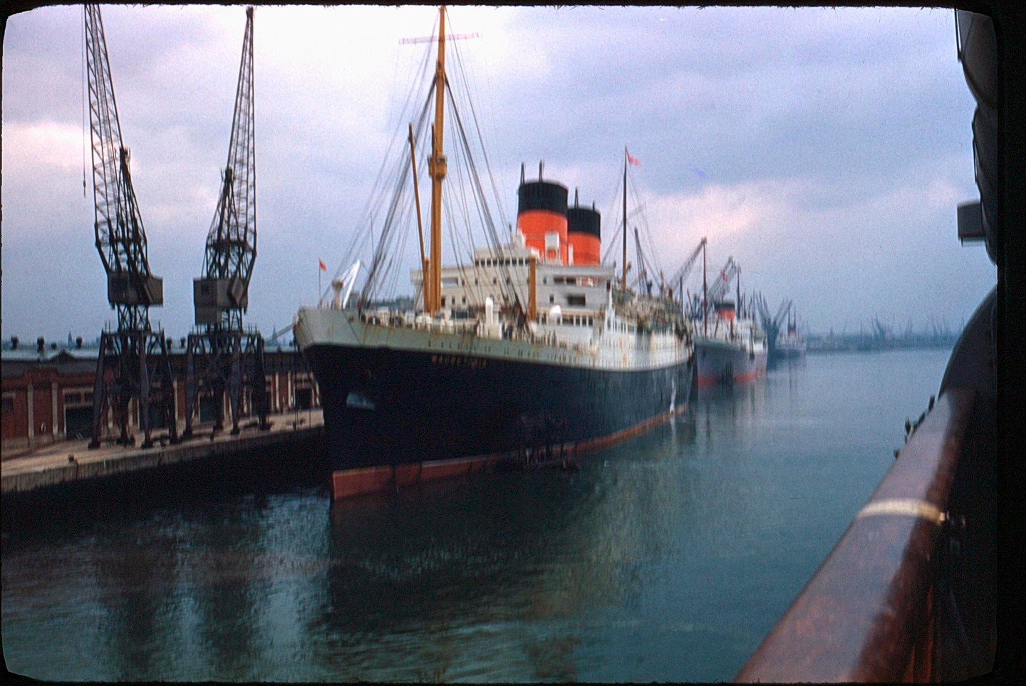 A large passenger ship with red and black smokestacks is docked next to a pier with cranes.