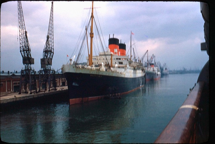 A large passenger ship with red and black smokestacks is docked next to a pier with cranes.