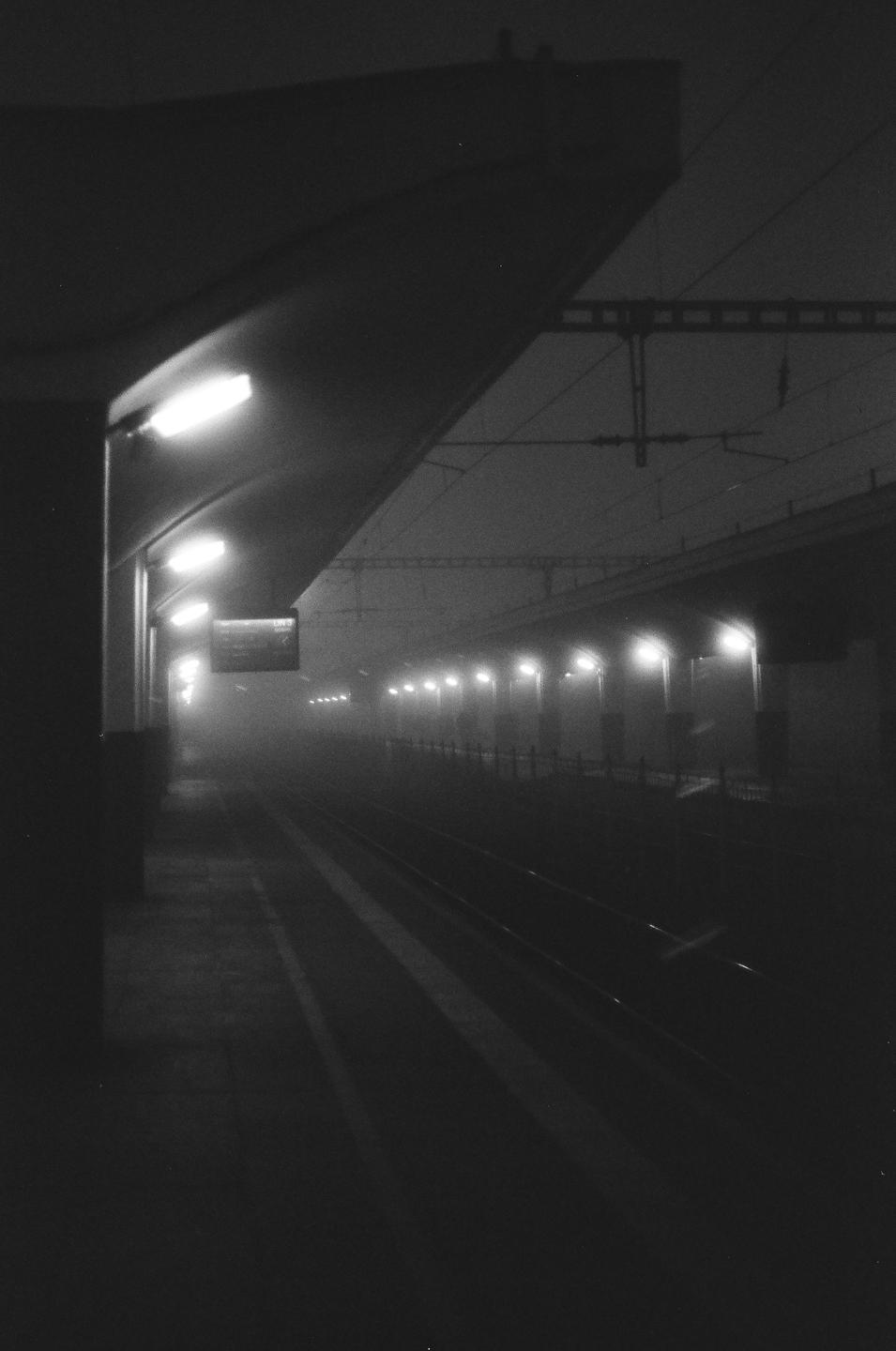 A train station platform is illuminated by overhead lights on a foggy night.