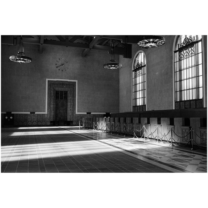 Sunlight streams through tall arched windows into a large, empty hall with a clock on the far wall.