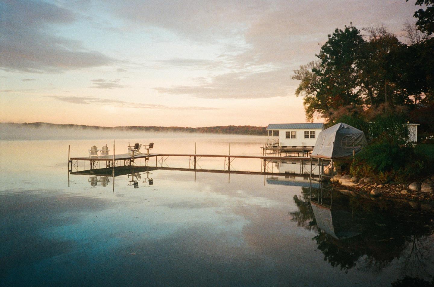 A long dock with chairs extends into a misty lake at sunrise, with a small house and tent on the shore.