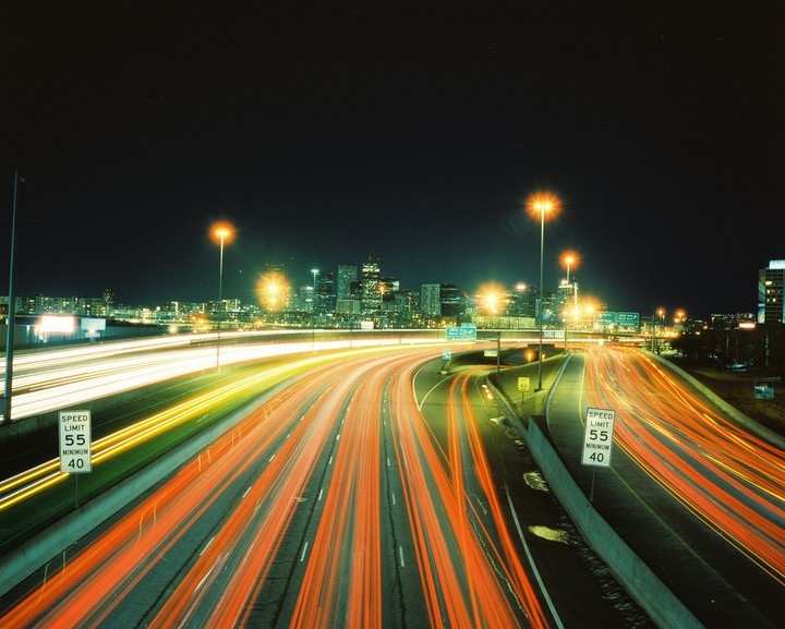 A highway at night shows streaks of light from vehicles with a city skyline in the background.