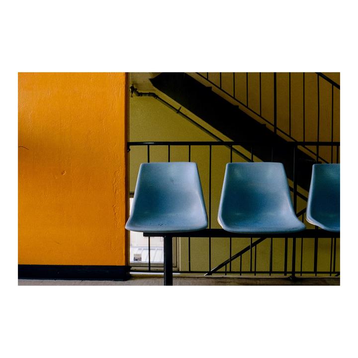 Three blue chairs are lined up against an orange wall next to a dark staircase.