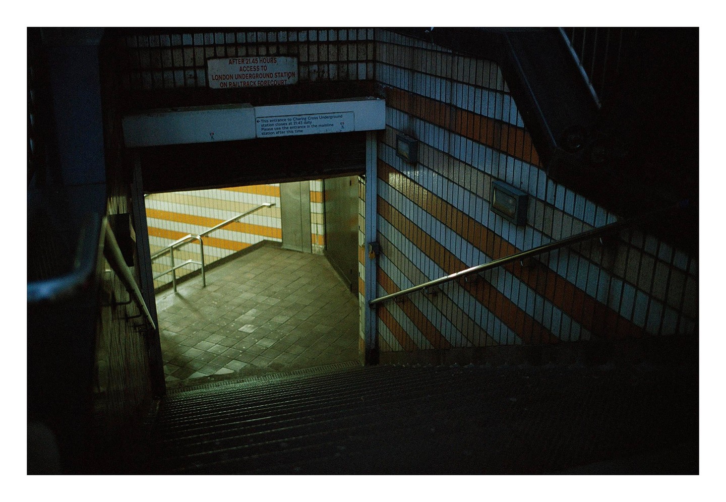 A dark stairwell leads down to a brightly lit underground station with striped walls.