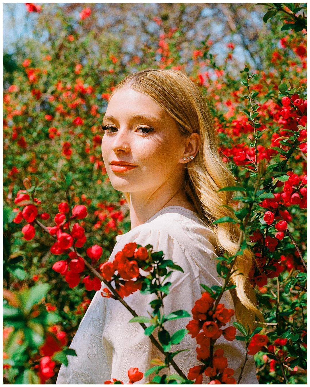 A woman with long blonde hair stands among red flowering bushes, looking over her shoulder at the viewer.