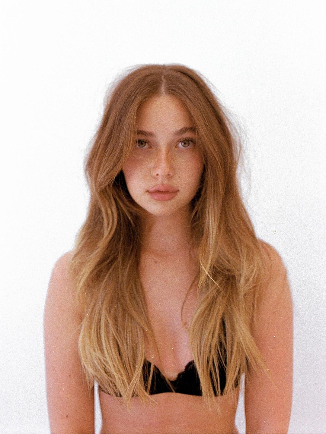 A woman with long wavy hair and a black bra stares directly at the viewer.