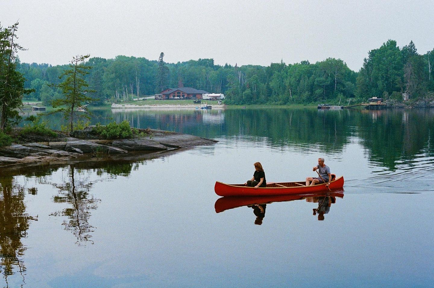 A couple paddles a red canoe on a calm lake surrounded by a forest and a distant building.