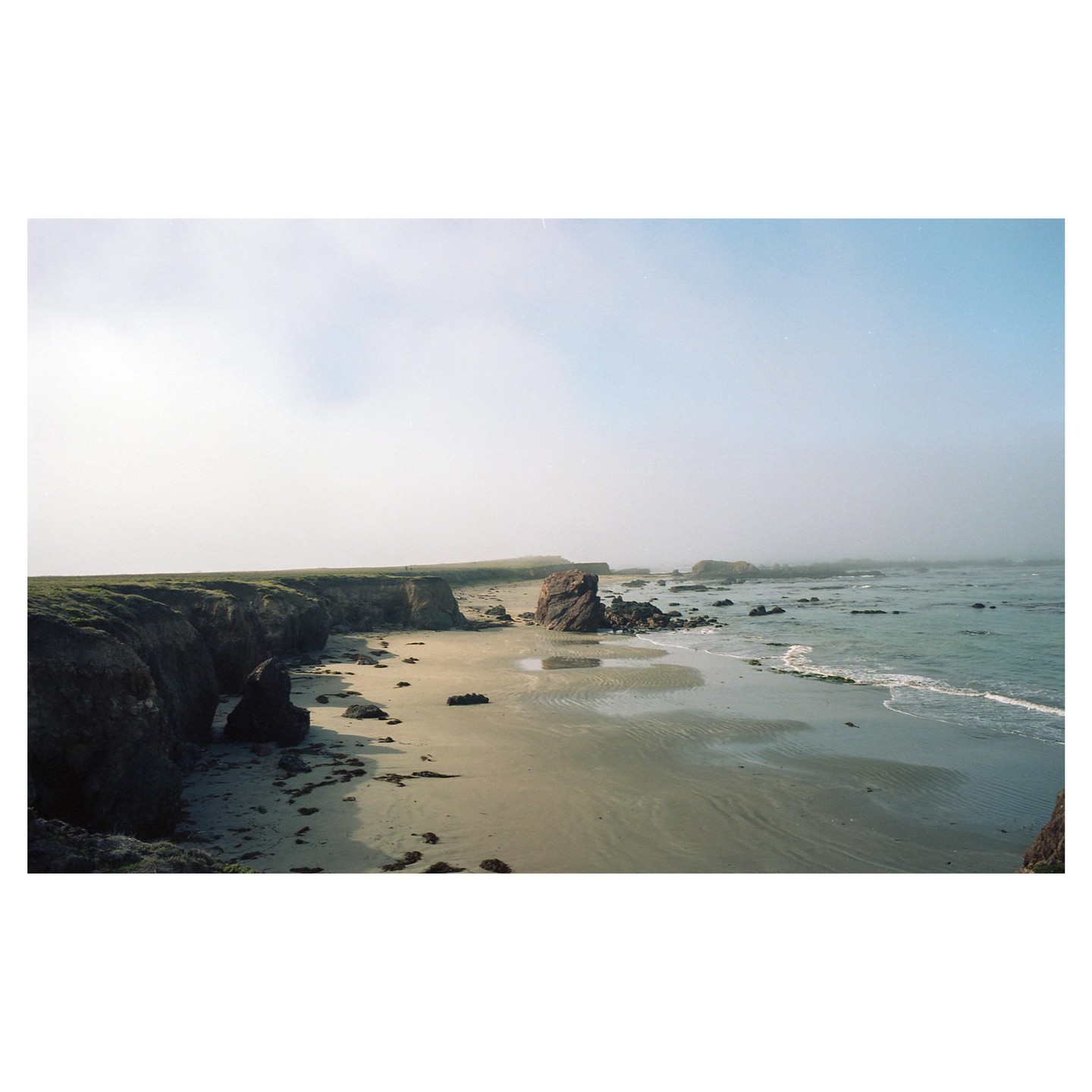 A sandy beach with cliffs and large rocks is partially obscured by fog.