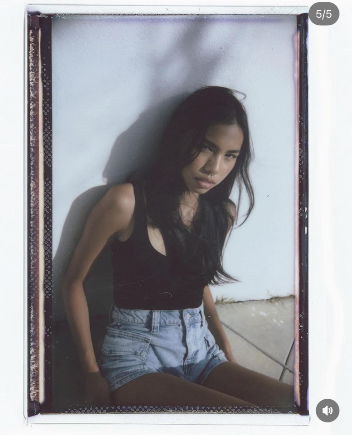 A woman with long dark hair and a nose ring sits against a white wall with shadows.