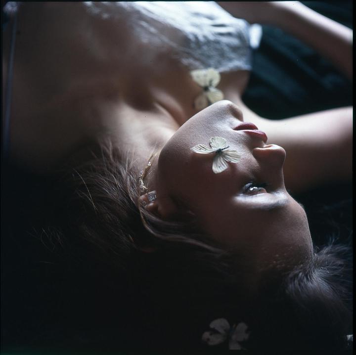 A woman lies on her back with a butterfly on her eyelid and another on her chest.