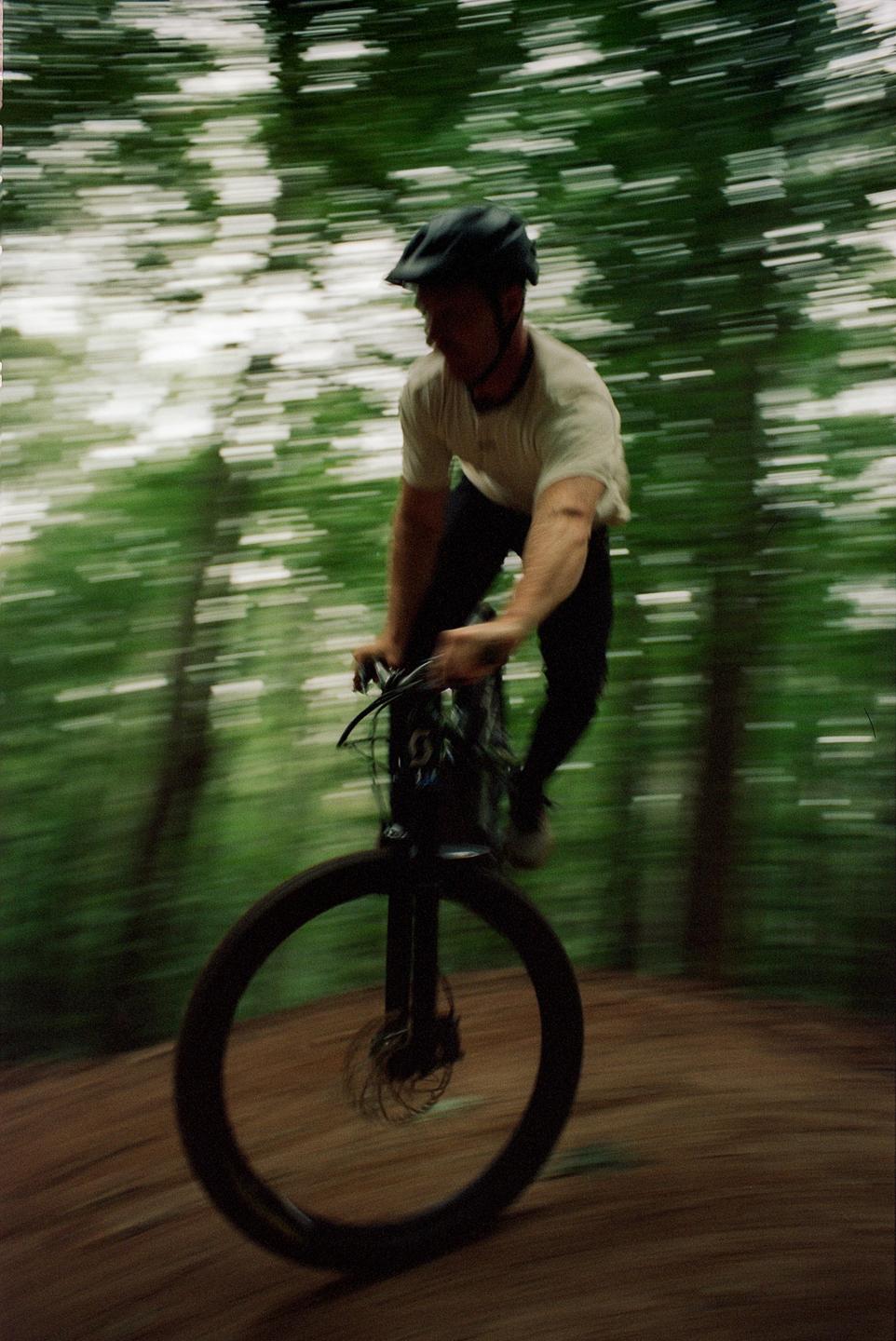A man on a bicycle rides through a forest with blurred trees in the background.