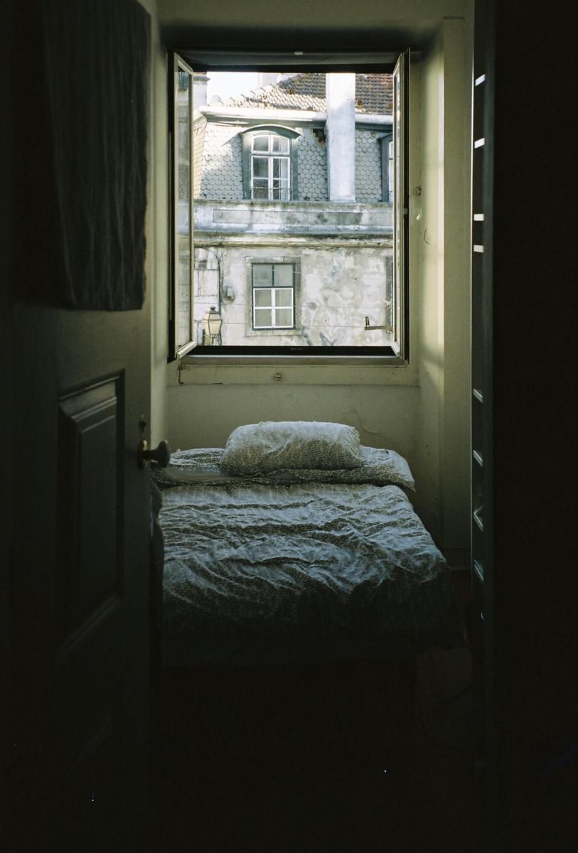 A bed with a patterned duvet and pillow is positioned beneath an open window looking out onto old buildings.