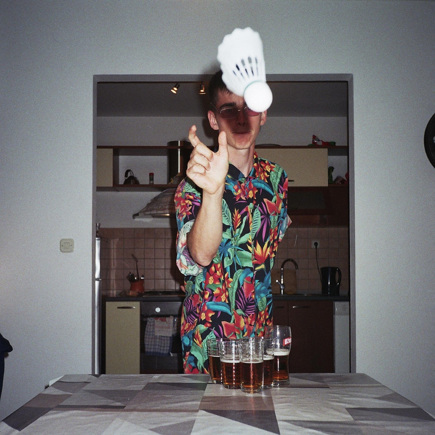 A man in a colorful shirt points at a shuttlecock floating above a table with several glasses of beer.