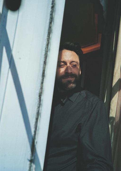 A man with a beard is looking out from behind a white door frame, with sunlight on his face.