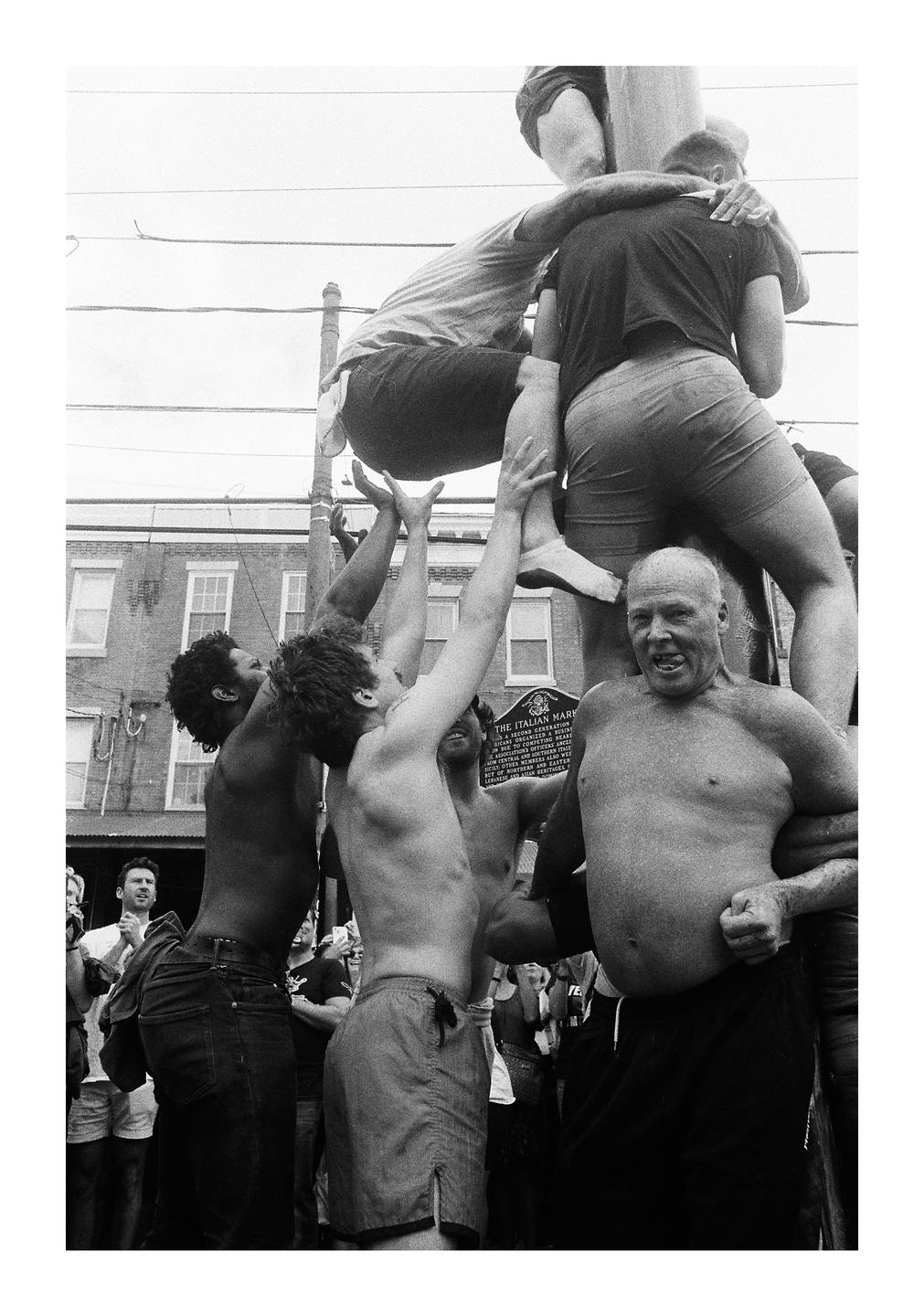 A group of men climb a greased pole in a street.