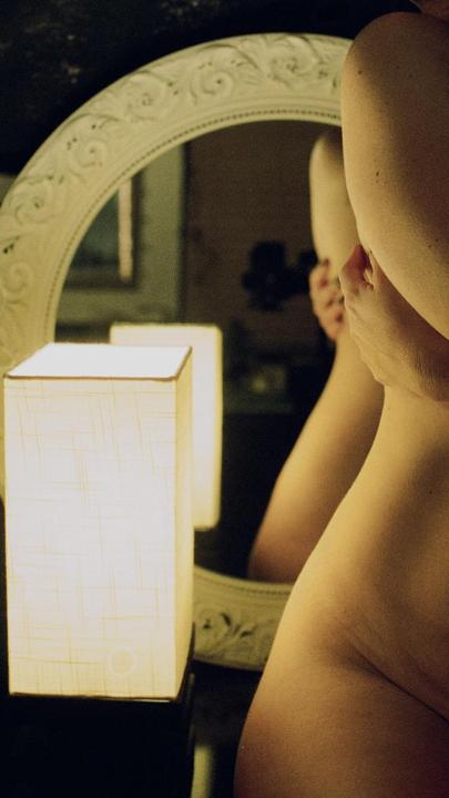A woman's torso is visible in the foreground, with her reflection in an ornate mirror behind a glowing lamp.