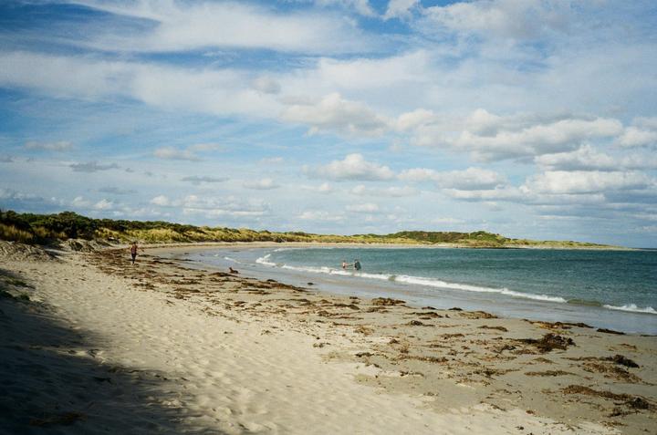 A sandy beach with a person walking along the shore and two people in the water under a cloudy sky.