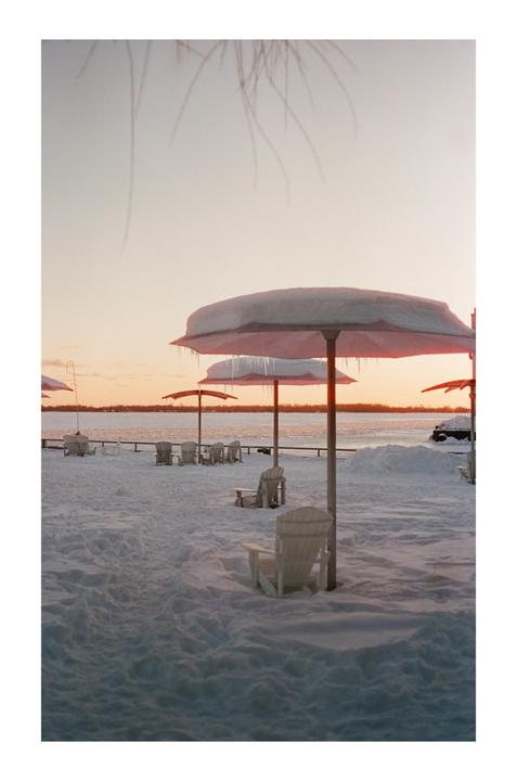 A snow-covered beach with umbrellas and chairs faces a body of water at sunset.