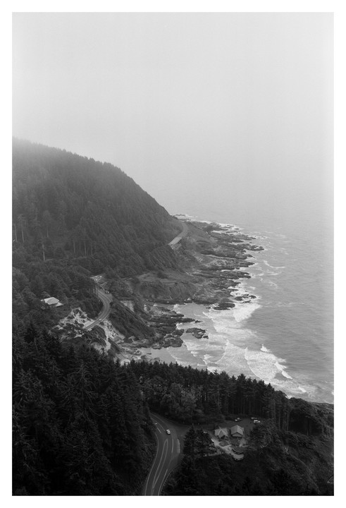 A winding road follows a forested coastline under a hazy sky.