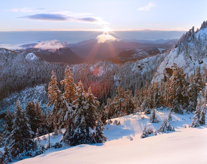 The sun rises over a snow-covered mountain landscape with evergreen trees.
