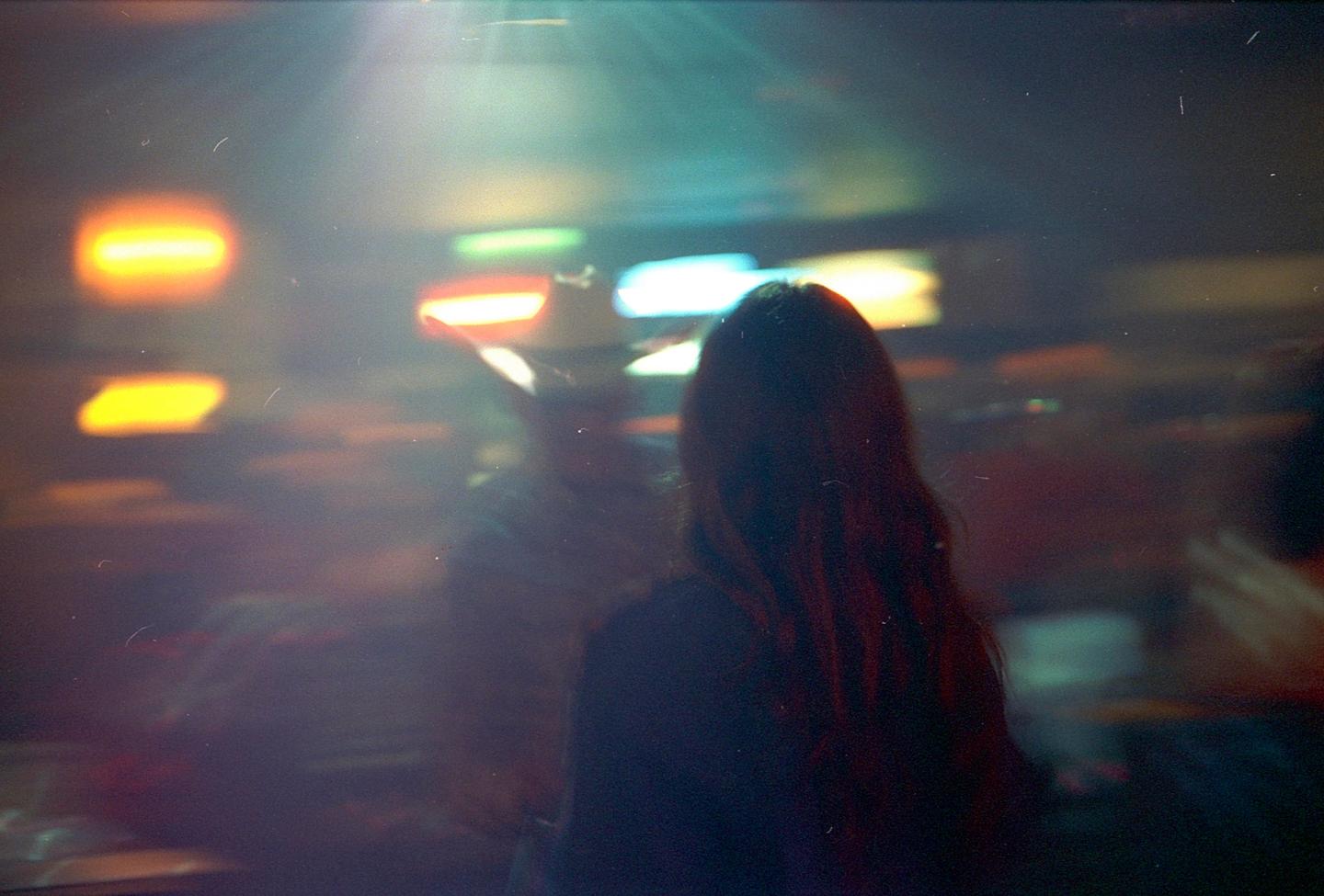 A woman with long hair stands in a dimly lit bar with colorful lights in the background.