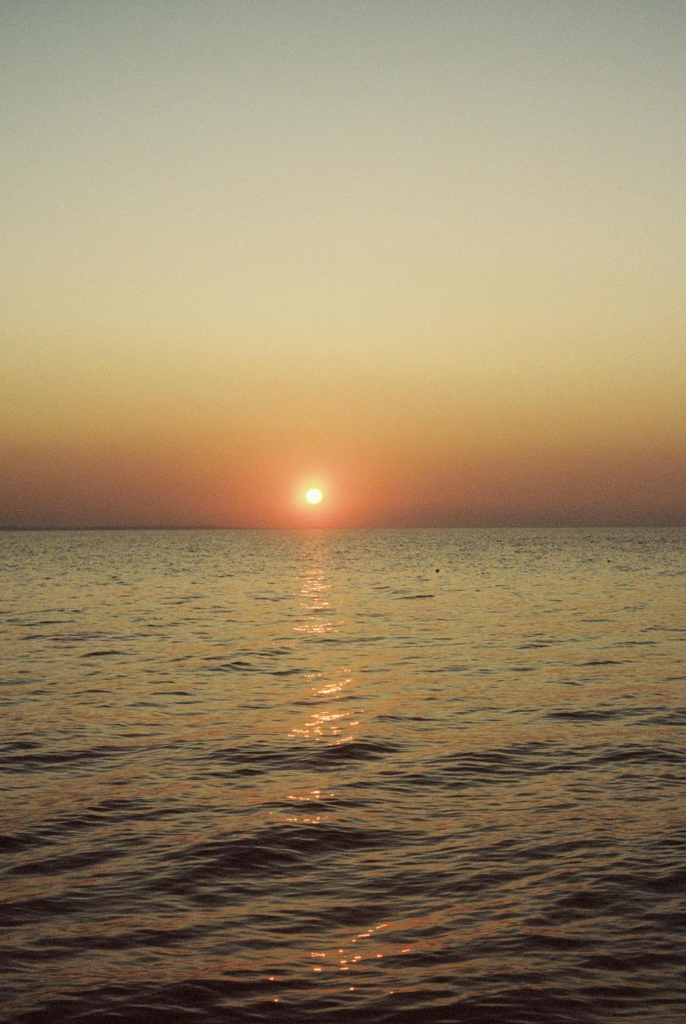 The sun sets over a calm body of water, casting a warm glow across the sky and reflecting on the ripples.