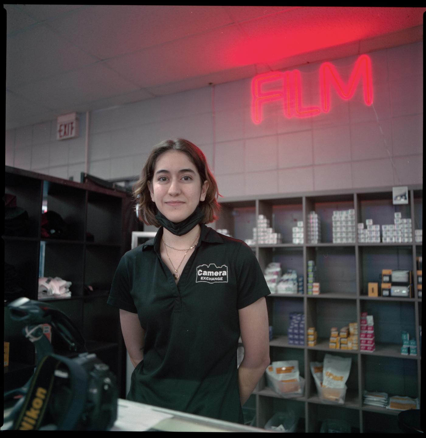 A woman wearing a black polo shirt with a camera store logo stands in front of shelves stocked with film and a red neon sign that reads "FILM."