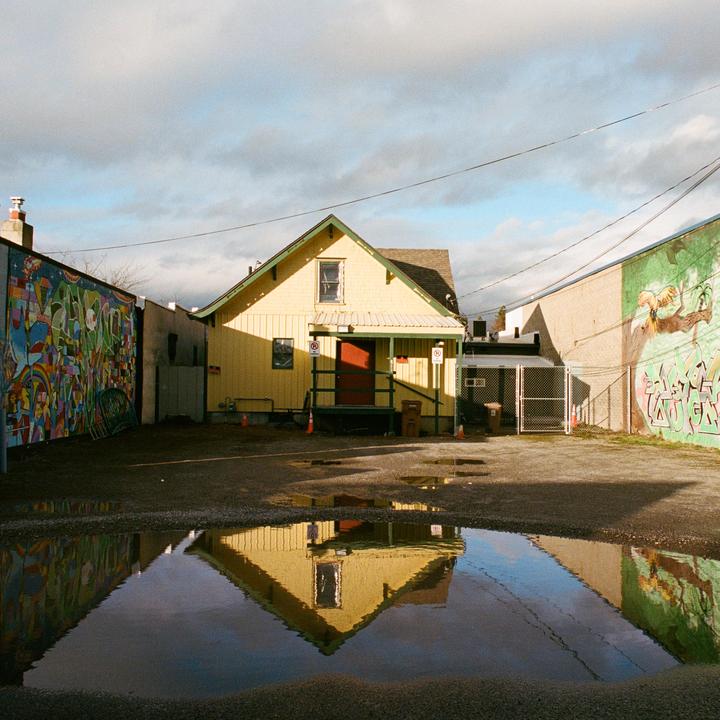 A yellow house with a red door is reflected in a puddle on a gravel lot between two buildings with murals.