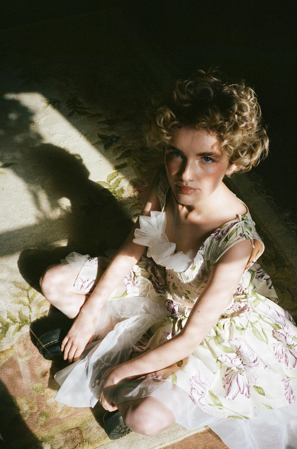 A young woman with curly hair and a floral dress sits on a patterned rug, looking up at the viewer.