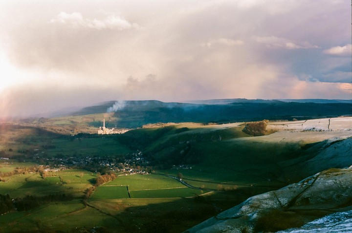 A valley with a town and factory is visible under a cloudy sky, with patches of snow on the hillsides.