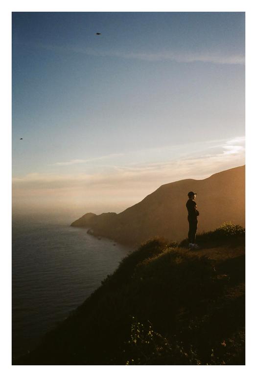 A man stands on a grassy cliff overlooking the ocean and mountains during golden hour.