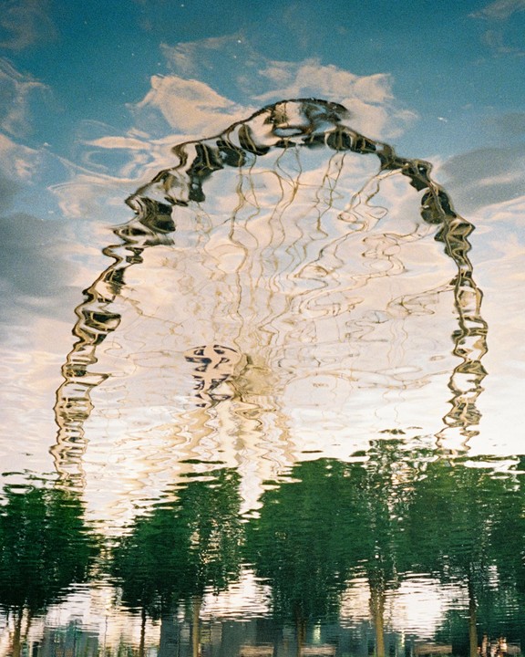 The London Eye and trees are reflected in rippling water under a blue sky with clouds.