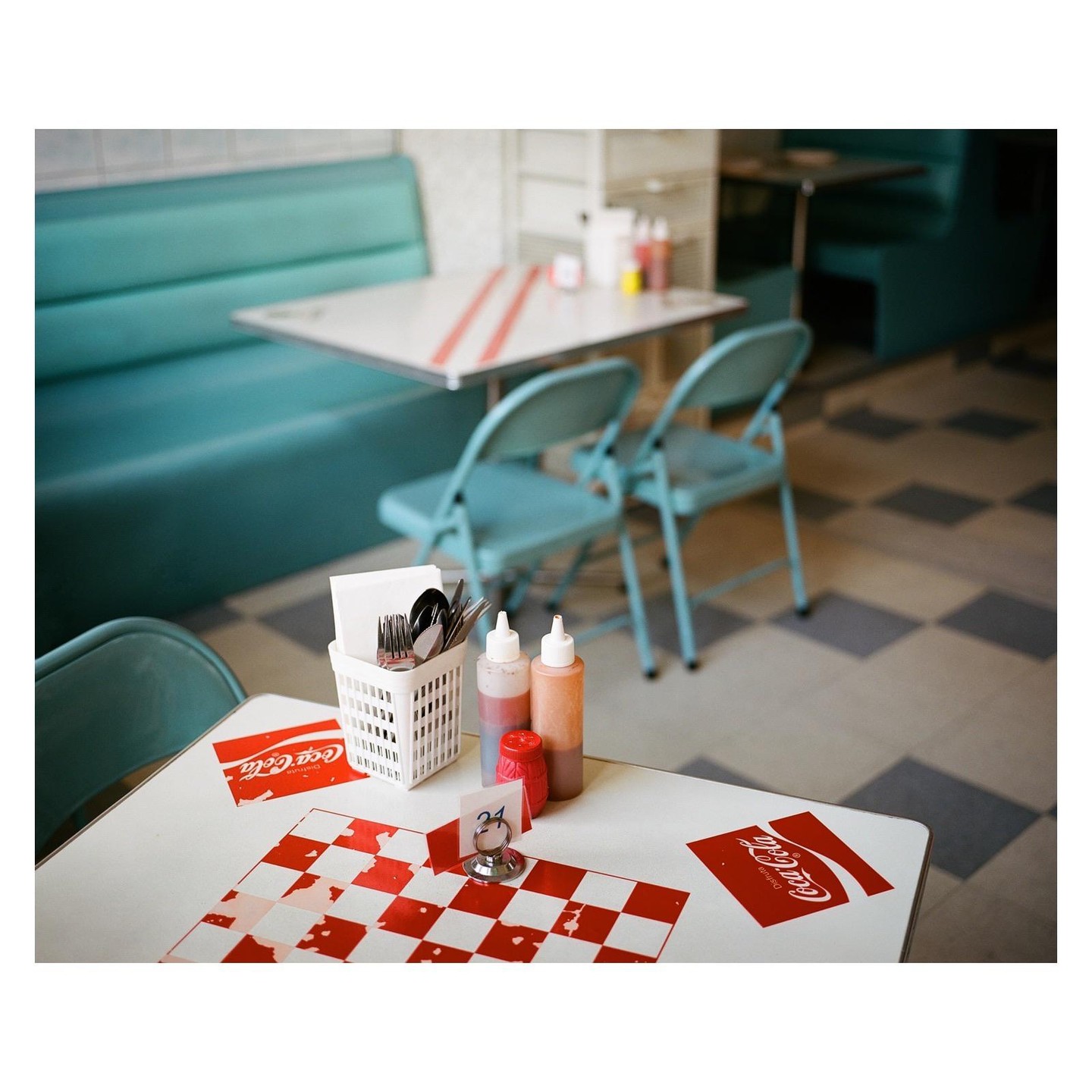 A diner table is set with condiments, cutlery, and a checkered placemat, with turquoise booths and chairs in the background.