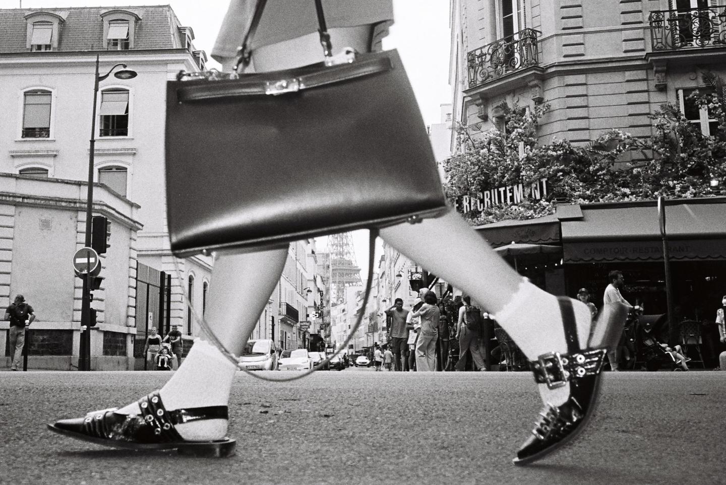 A low-angle street shot shows a woman's legs and handbag as she walks down a street in Paris, with the Eiffel Tower visible in the distance.