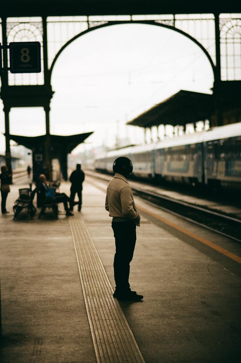 A person wearing headphones stands on a train platform with a train on the tracks and other people in the background.