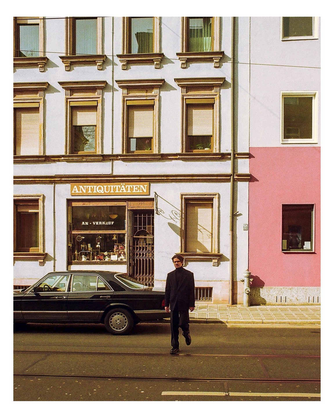 A man in a dark suit walks across a street in front of an antique shop and a parked car.