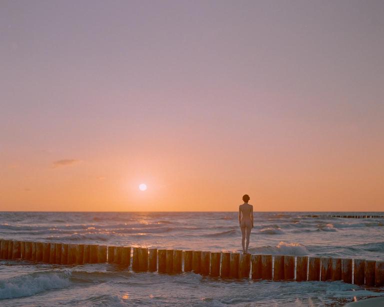 A woman stands on a wooden pier looking out at the ocean during sunset.