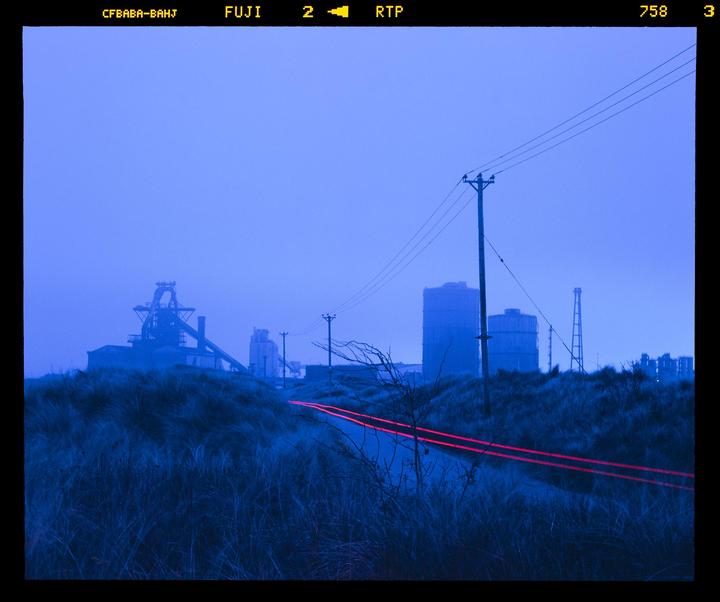 Red light trails from a vehicle on a road at dusk with industrial buildings in the background.