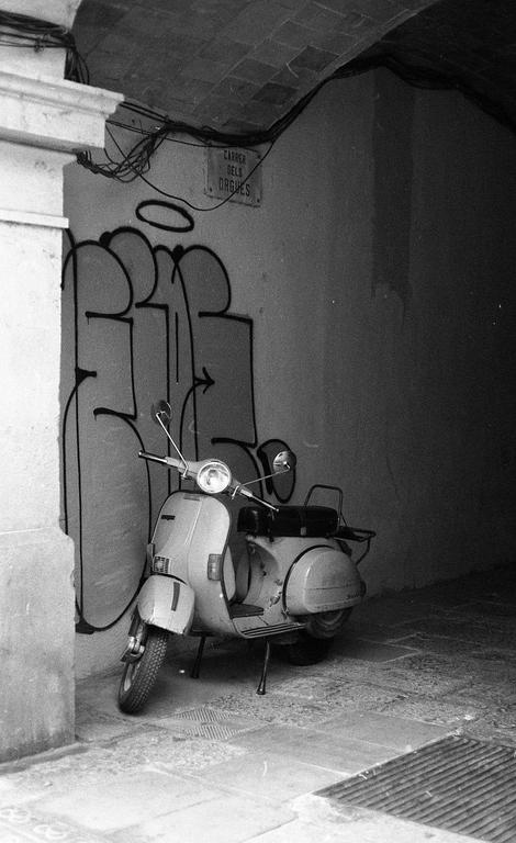 A scooter is parked next to a wall with graffiti and a street sign in a narrow alley.