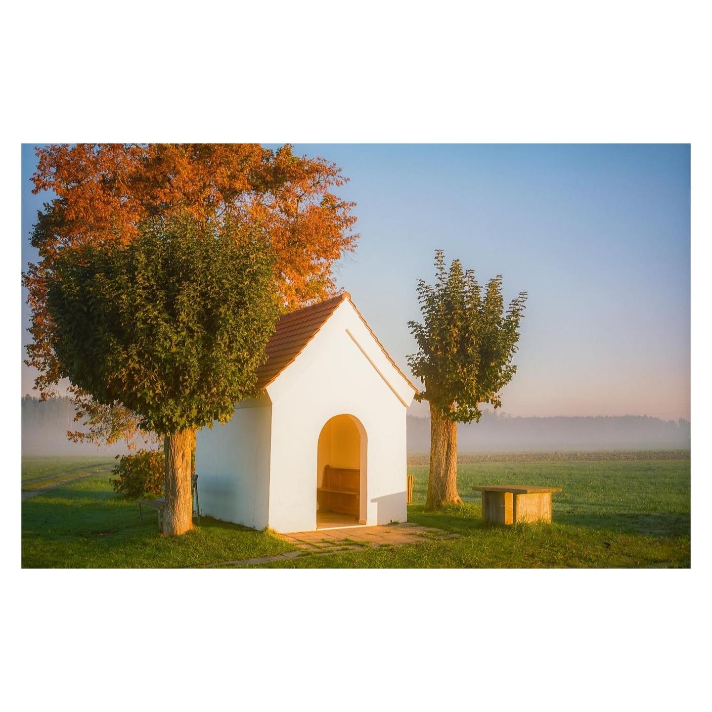 A small white chapel with a red roof stands between two trees in a green field under a clear sky.