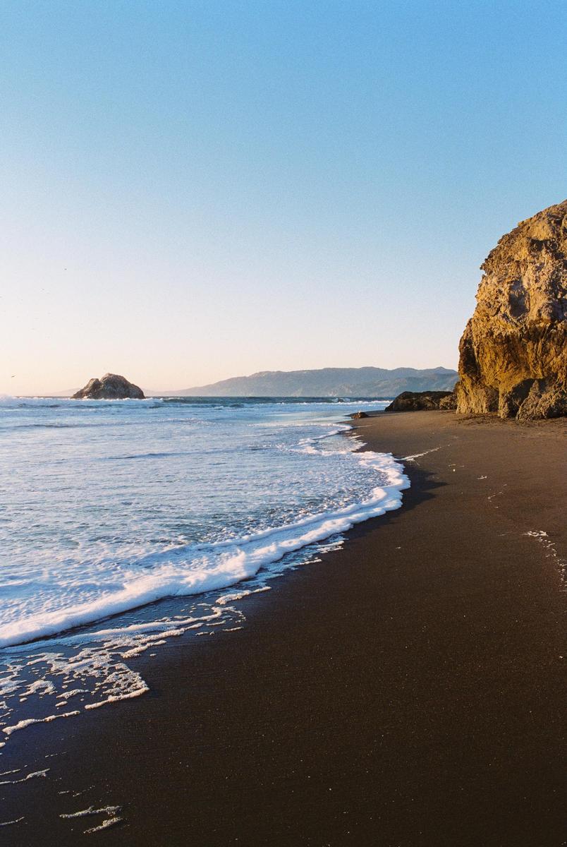 Waves crash on a dark sand beach next to a large rock formation under a clear sky.